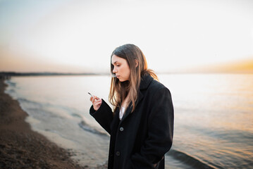 Sad young woman 24-26 year old wearing black winter coat standing at beach over sea and sunset...