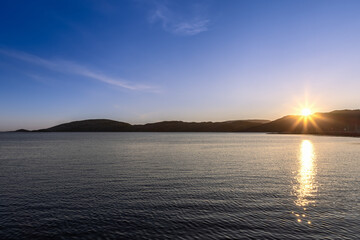 Midnight summer sun peeking out from behind a hill, illuminating the fjord near the port of Bodo on...