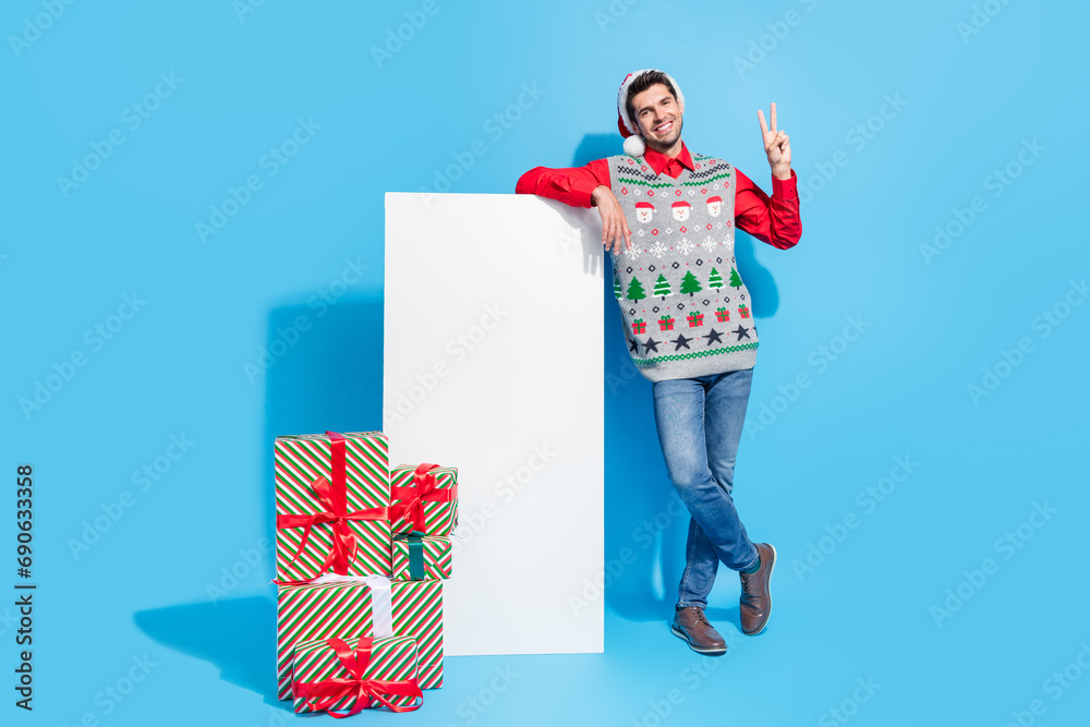 Poster Full size photo of handsome young guy vertical banner v-sign presents wear x-mas print vest hat outfit isolated on blue color background