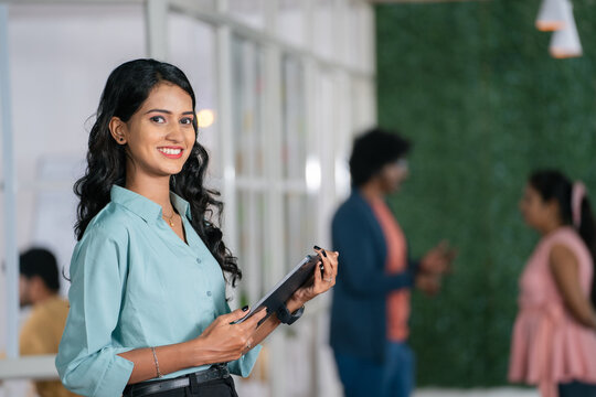 confident young woman employee standing at office by looking camera with tab in hand - concept of businesswoman, startup entrepreneur and career growth