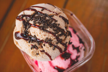 Imagen horizontal y cenital de un tazón de vidrio con bolas de helado de chocolate o café y fresas con crema postre de verano sobre una mesa de madera