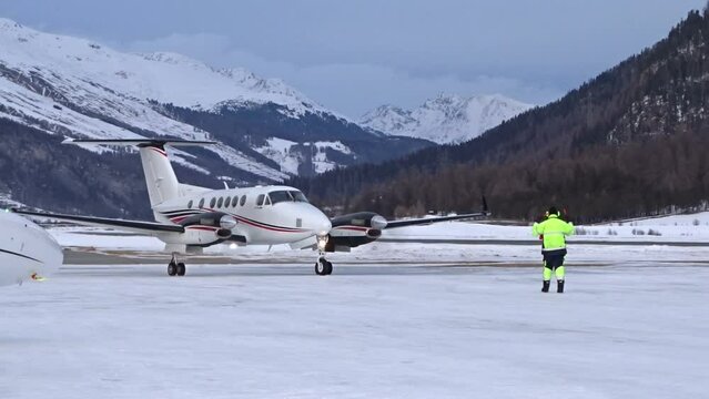 Close Up Twin engine turboprop aircraft arriving at executive airport with marshaller signing signaling guiding airplane in winter landscape cold area mountains valley skiing resort