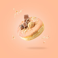 Sweet donut with pink coral icing, chocolate pieces, candy and sprinkles flying isolated on gradient peach fuzz background.