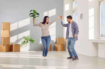 Happy young couple celebrating moving day with cardboard boxes in background smiling and dancing with plant in hands in new appartment. Family having fun. Relocating, real estate, mortgage concept.