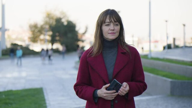 An attractive middle-aged Caucasian woman thinks about something as she holds a smartphone in an urban area
