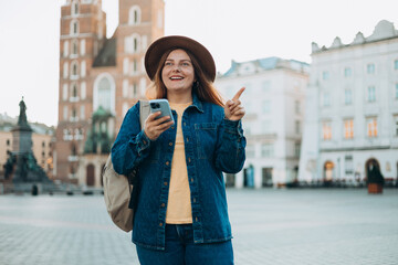 Attractive young female tourist is exploring new city. Redhead girl pointing finger and holding a smart phone on Market Square in Krakow. Traveling in summer. St. Marys Basilica. Vacation concept