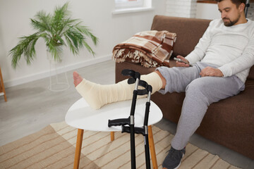 Man with a broken leg using a modern cellphone while sitting on the sofa at home, with his foot resting on a stool, with orthopedic crutches beside. Injury, accident, treatment concept - obrazy, fototapety, plakaty