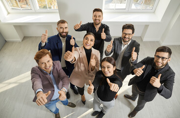 Team of happy, satisfied business people giving thumbs up all together. Joyful young men and women standing in office, looking up, smiling and doing like gestures. Shot from above. Success concept - obrazy, fototapety, plakaty