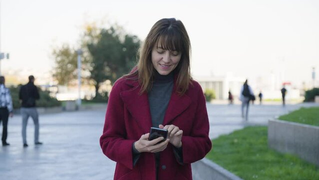 An attractive middle-aged Caucasian woman works on a smartphone with a smile as she walks in an urban area