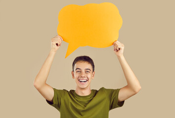 Happy excited young boy wearing green t-shirt holding blank speech bubble for thoughts isolated on beige studio background. Guy with empty word cloud offering space for text. Idea concept. - obrazy, fototapety, plakaty