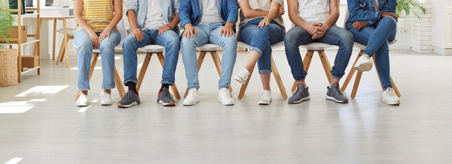 Diverse group of different young people wearing casual shirts, modern blue jeans and white and gray sneakers sitting on row of chairs in office workplace. Cropped low section shot of human legs - Powered by Adobe