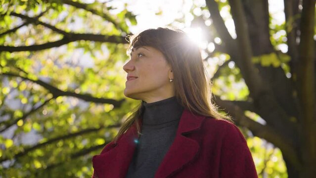 An attractive middle-aged Caucasian woman looks around and smiles in a park - closeup from below - behind her, sun shines through a tree crown