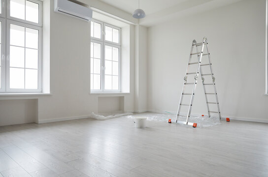 Empty unfurnished room with ladder and paint bucket. Unfinished living room interior with white walls, floor and windows. Renovation. Repairs in apartment. Empty interior space