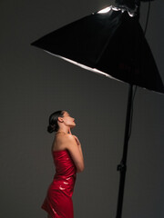 Young pretty woman poses in red latex dress under spotlight in studio.