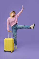 Happy blonde girl tourist holding travel suitcase standing isolated on purple background. Smiling...