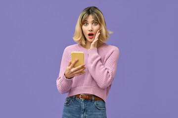 Shocked young gen z blonde woman shopper holding smartphone using mobile cell phone surprised about wow fashion sale app, omg promo discounts on cellphone isolated on purple background. - obrazy, fototapety, plakaty