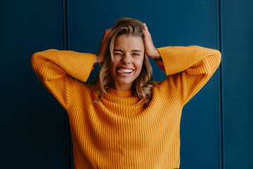 Playful young blond hair woman in yellow sweater sticking out tongue while having fun on blue wall