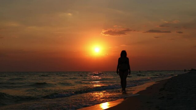 A woman walks along the seashore with the surf and sunset in the background. Slow Motion