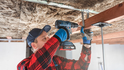 Lowering and insulate the ceiling. Fixing a wooden prism using tin hanger to the load-bearing wooden structure of the ceiling. The worker works with a cordless drill.