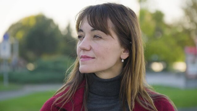 An attractive middle-aged Caucasian woman looks into distance with a melancholic smile as she stands in a park - closeup