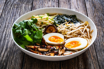 Asian style soup. Broth with chow mein noodles, boiled eggs, shiitake mushrooms, cilantro and mung sprouts on wooden table

