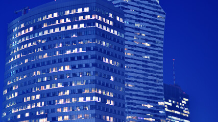 Fototapeta na wymiar Fragment of the glass facade of a modern corporate building at night. Big glowing windows in modern office buildings at night, in rows of windows light shines. Blue graphic filter.