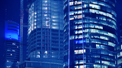 Fototapeta na wymiar Fragment of the glass facade of a modern corporate building at night. Big glowing windows in modern office buildings at night, in rows of windows light shines. Blue graphic filter.
