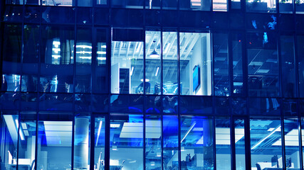 Fragment of the glass facade of a modern corporate building at night. Big glowing windows in modern office buildings at night, in rows of windows light shines. Blue graphic filter.