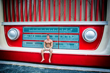 A young shirtless man sitting on a bench in front of a giant radio