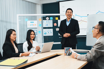 Successful businessmen present financial projects on glass whiteboard confidently. Executive managers listen to employee's quarterly business plan presentation. Modern meeting room. Intellectual.