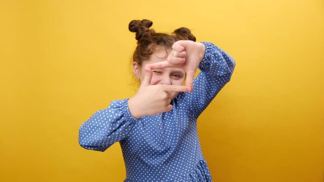 Portrait of cute little girl kid making frame with fingers and smiling looking at camera, pretending to make photography, posing isolated over yellow color background wall in studio with copy space
