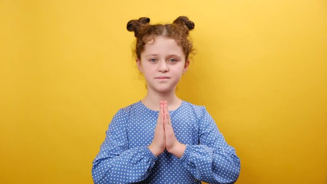 Sincere kids faith. Portrait of cute peaceful little girl praying to God, making prayer gesture, happy looking at camera, posing isolated over yellow color background wall in studio. Religion concept