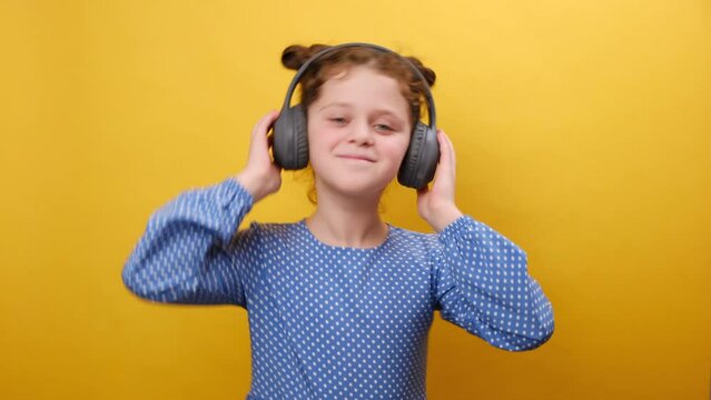 Happy mood concept. Portrait of cheerful pretty little girl listening to music in big wireless headphones and dancing, happy looking to camera, posing isolated on yellow background wall in studio
