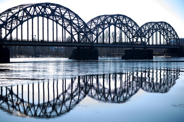 
A metal arched bridge across the river with the reflection of the bridge in the water in the form...