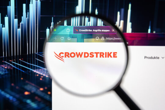 Crowdstrike company logo on a website with blurry stock market developments in the background, seen on a computer screen through a magnifying glass.	