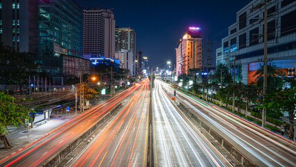 Nighttime time-lapse image of a large, four-lane, straight, parallel concrete road. It is flanked by a two-lane road. Surrounded by tall buildings, many white roadside lights, a bus stop,