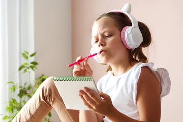 An 11-year-old blonde girl holds a notebook in her hands, headphones on her head, looking thoughtfully to the side
