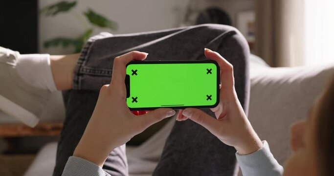 Happy young woman using smartphone with mockup green screen in horizontal landscape mode, resting on sofa couch. Watching movies on phone. Advertisement of app. Watching content, videos, blogs. POV