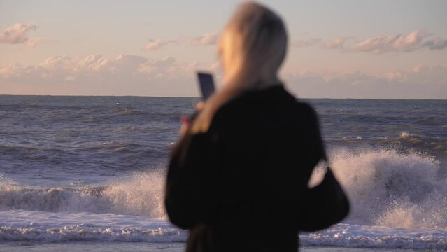 Back view of woman in coat standing in front of sea. Filming sea with mobile phone camera. Sad and emotional man looking into distance. Ship on horizon.