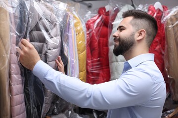 Dry-cleaning service. Happy worker choosing clothes from rack indoors