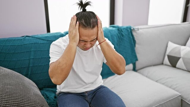 Stressed young chinese man, frustrated and alone, sits on sofa nursing a headache at home amidst sadness and personal conflict