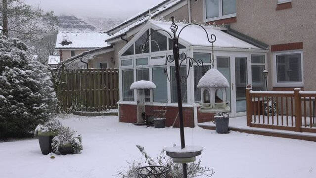 Small garden birds eating seeds from a snow topped garden feeder. In the background is a snow-covered conservatory and in the distance are snow-capped hills.