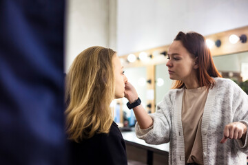 Make up artist woman doing perfect makeup young lady in beauty salon at mirror backdrop. Customer...