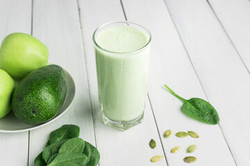 Green detox smoothie, blended vegetarian drink in a glass from spinach, apple, avocado and seeds on white wooden table, healthy eating concept