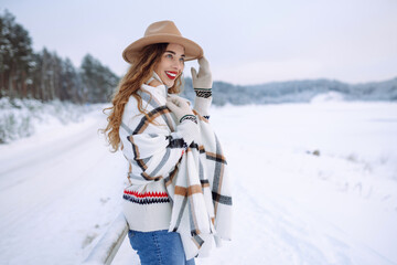 Fototapeta na wymiar Portrait of a smiling woman in a hat and scarf in a snowy forest enjoying a winter day. Young traveler posing outdoors. Adventure, weekend concept.