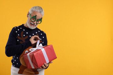 Senior man in Christmas sweater and funny glasses opening gift against orange background. Space for text
