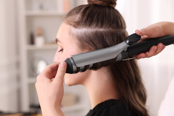 Hairdresser curling woman's hair with iron in salon, selective focus
