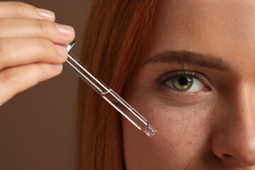 Beautiful young woman applying cosmetic serum onto her face on brown background, closeup