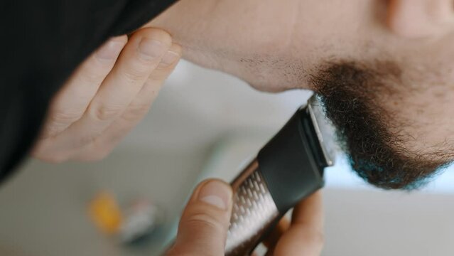 
Close-up of a man shaving his beard with an electric trimmer, focusing on the tool in hand and the detail of the facial hair.