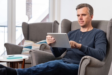 handsome 40s single man sits in an comfy  grey armchair and uses tablet in landscape mode
